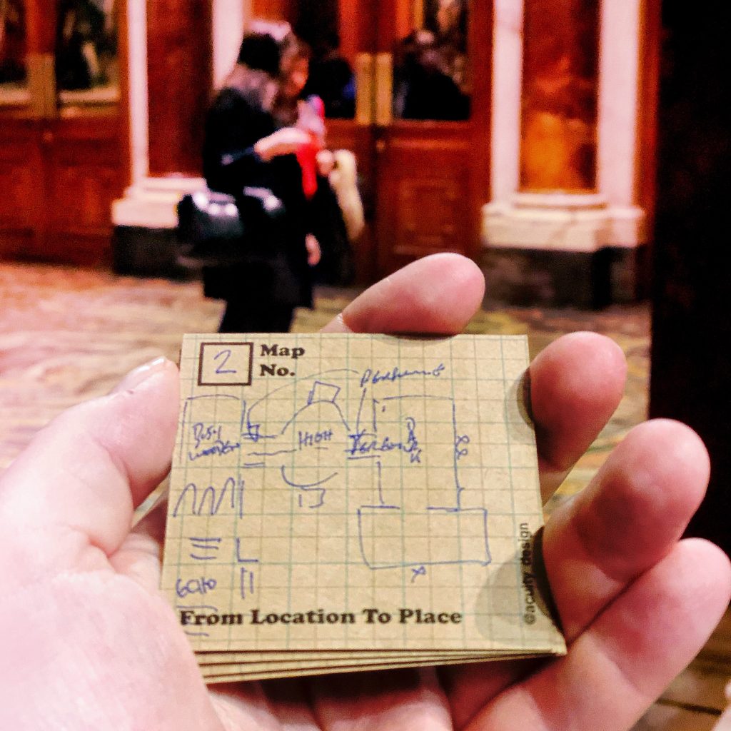 Square postcard held in had with map sketched on it. In the background are people walking thru the museum.